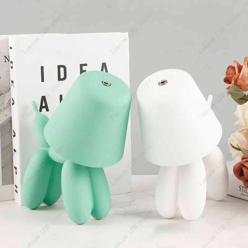 Two plush toys, one green and one white, on a light gray surface with a book and flowers in the background.
