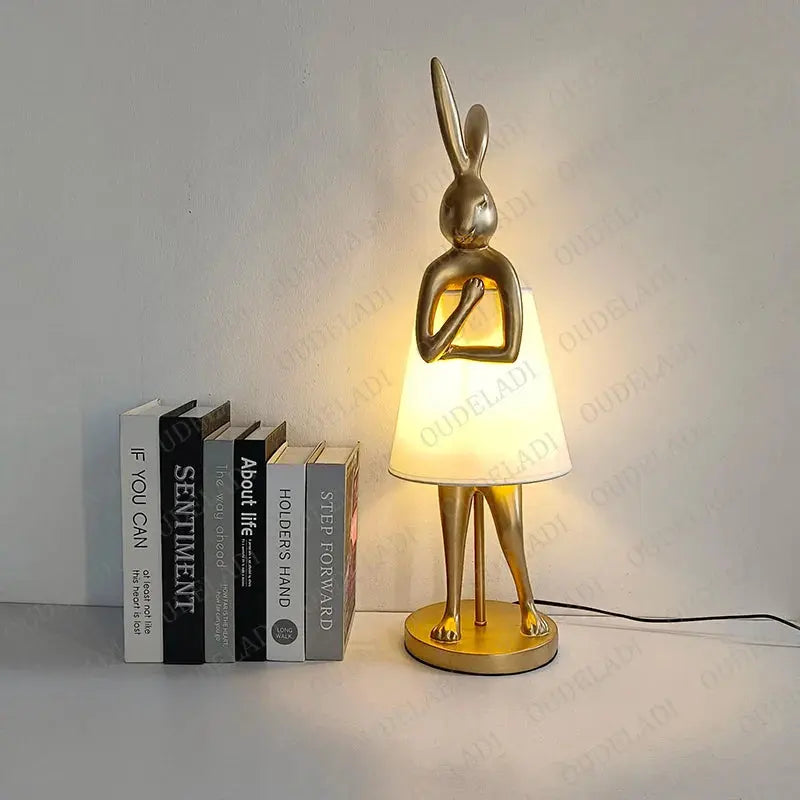 Gold rabbit-shaped lamp with a white shade on a gray surface next to books.