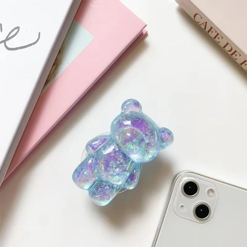 Iridescent bear-shaped phone case on a white surface with a phone and books in the background.