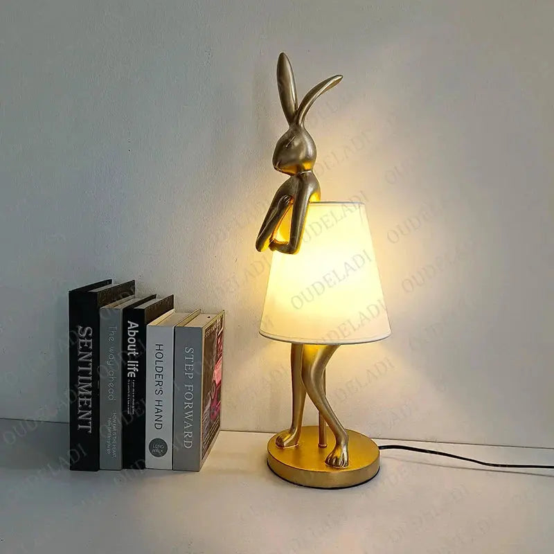 Gold rabbit-shaped table lamp with a white shade on a surface next to books.