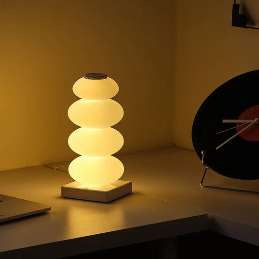 Stacked cylindrical lamp on a desk with a laptop and clock in the background