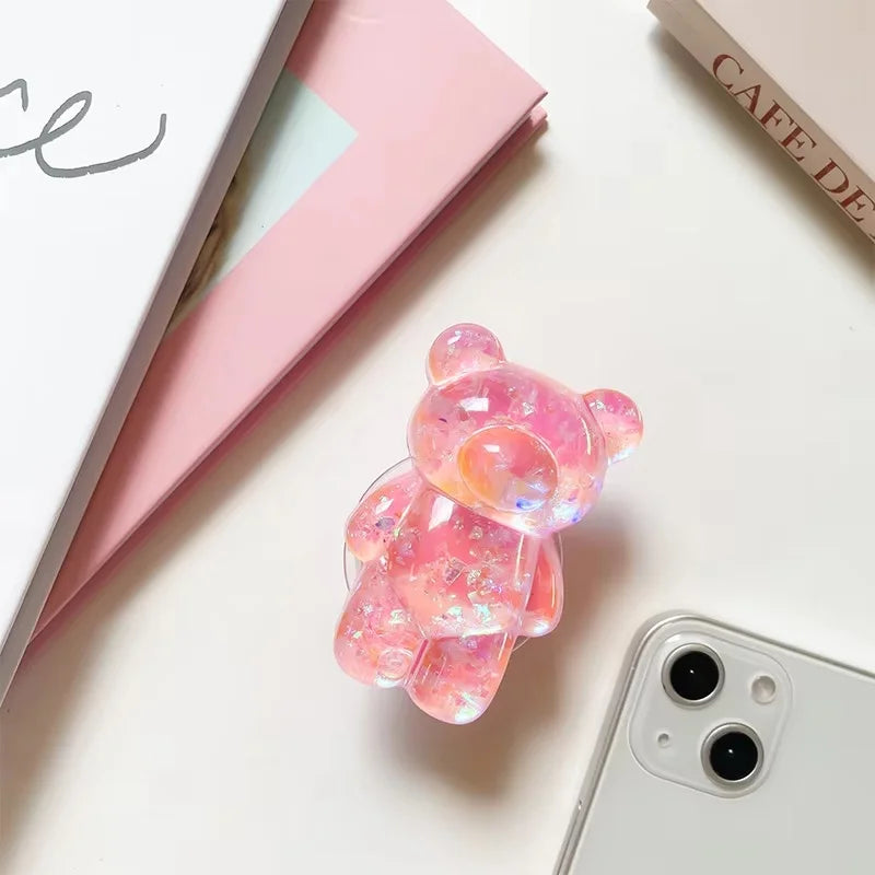 Pink bear-shaped phone case on a light surface with a phone and notebook in the background