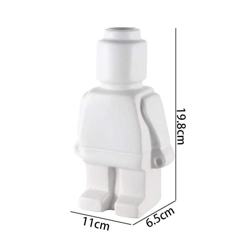 Two figurine vases shaped like Lego men, one black and one white, with the black one displayed in front of the white one. The black vase contains a dry branch, and the white vase has a small green plant.