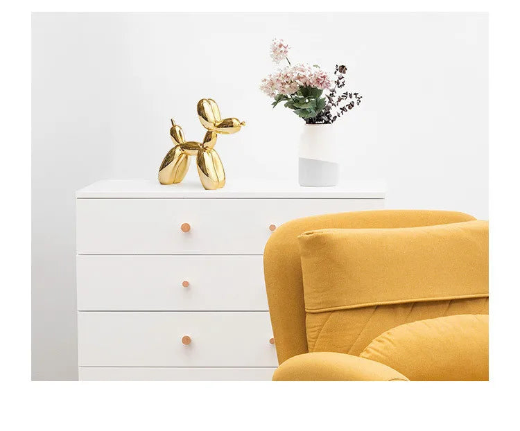 White dresser with gold dog sculpture and white vase with flowers, next to a yellow armchair.