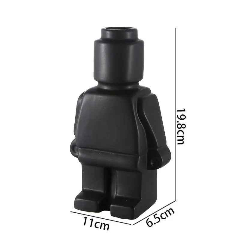 Two figurine vases shaped like Lego men, one black and one white, with the black one displayed in front of the white one. The black vase contains a dry branch, and the white vase has a small green plant.