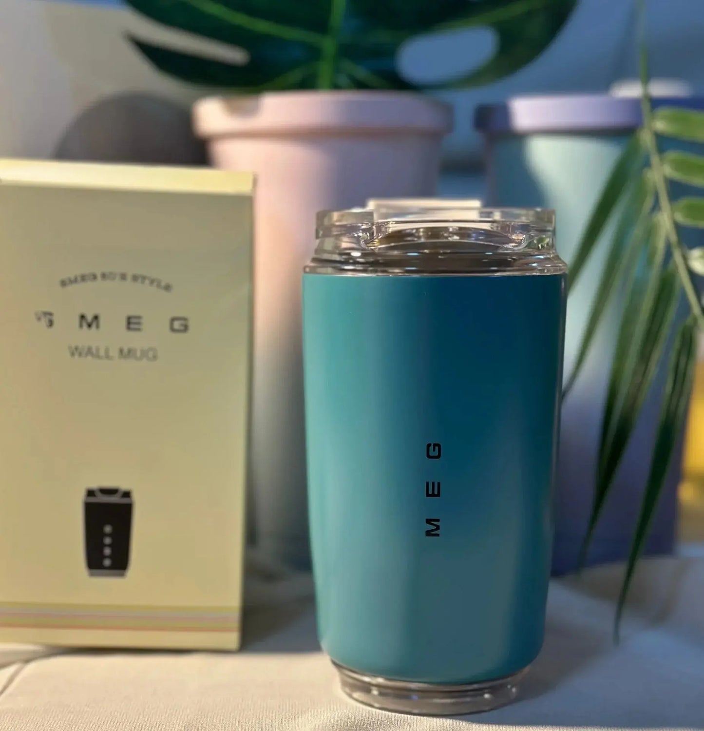 SMEG Blue mug with 'SMEG' branding next to a box and plant