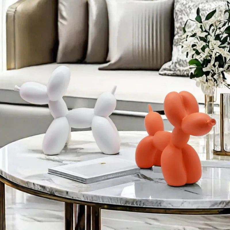 Two balloon dog sculptures, one white and one orange, on a marble coffee table with a couch and decorative items in the background.