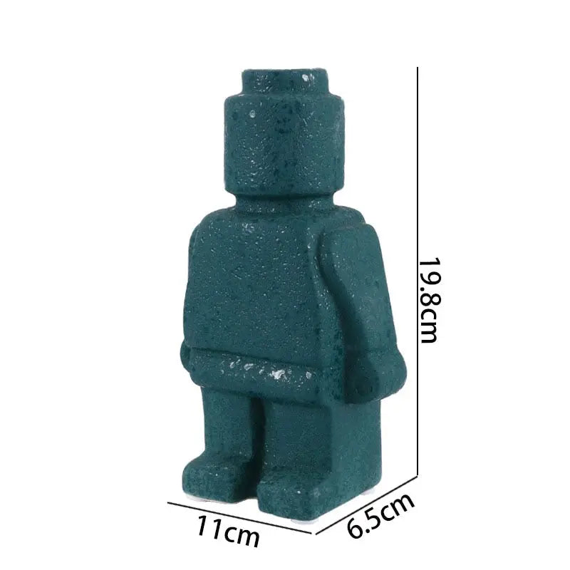 Two figurine vases shaped like Lego men, one black and one white, with the black one displayed in front of the white one. The black vase contains a dry branch, and the white vase has a small green plant.