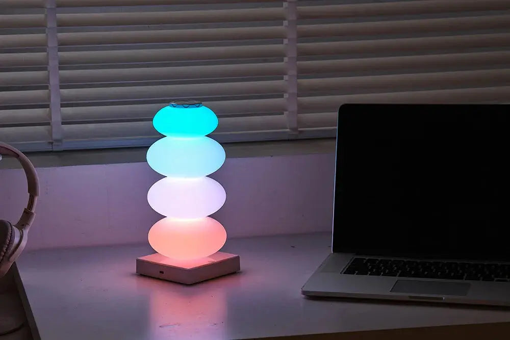 Colorful lamp on a desk next to a laptop with blinds in the background