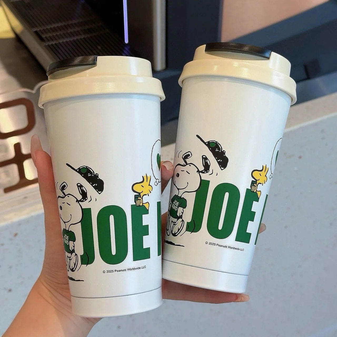 Two white coffee cups with cartoon characters and 'JOE' text held in front of a coffee machine.