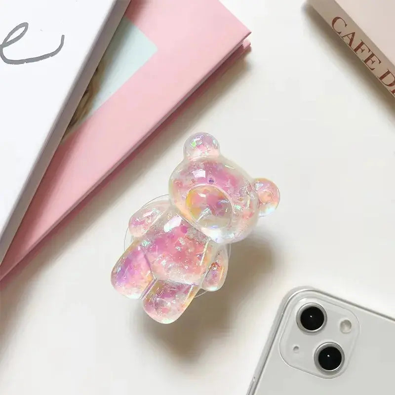 Iridescent bear-shaped phone case on a white surface with a pink notebook and phone.