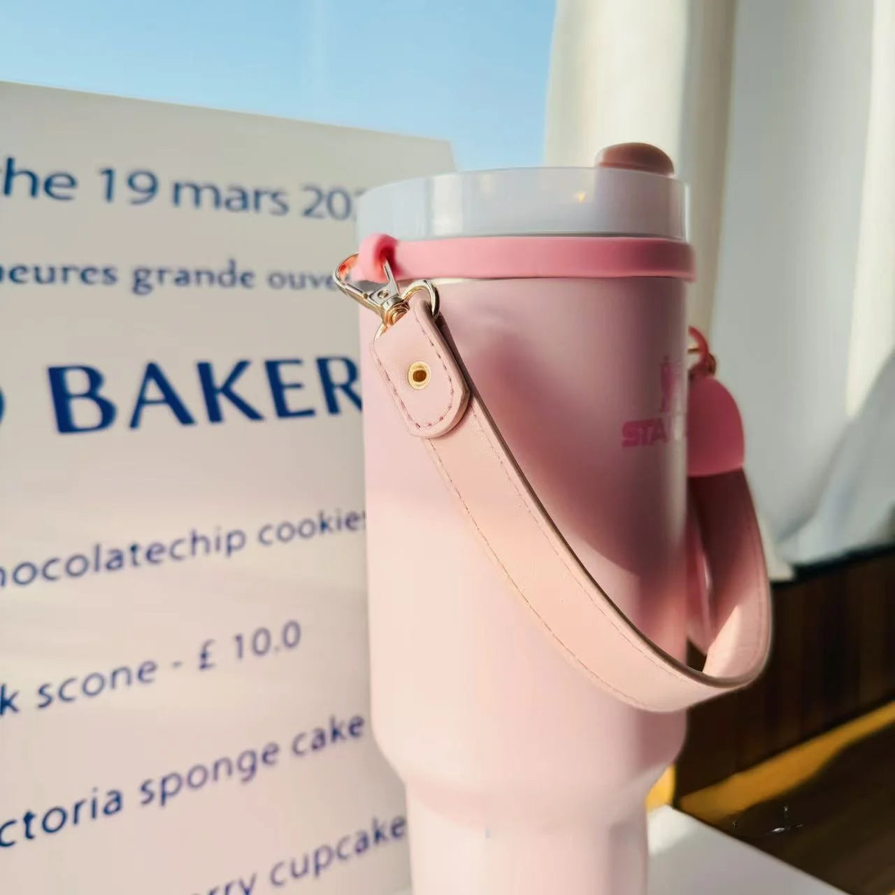 Pink water bottle with a strap held by a hand, blurred background with text and a window.