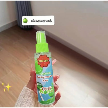 Hand holding a clothing fragrance spray labeled 'crispy green apple' on a wooden floor.