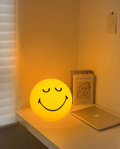 Smiley face lamp on a desk next to a laptop and book