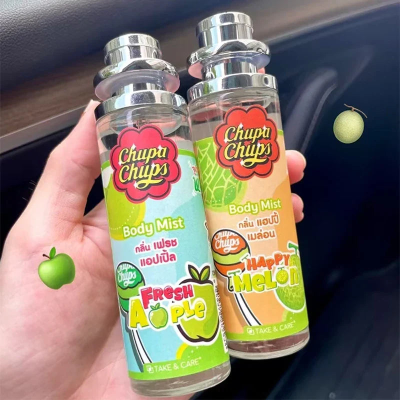 Two Chupa Chups body mist bottles, one 'Fresh Apple' and one 'Happy Melon', held in a hand with a blurred background.