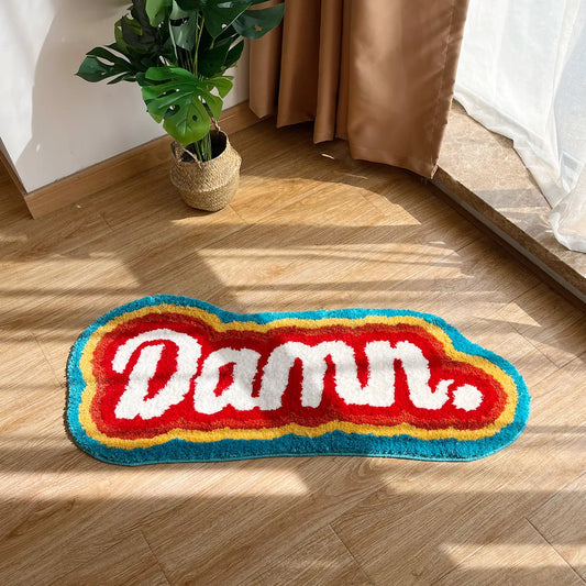 Colorful rug with 'Damn.' text on a wooden floor, next to a plant and curtain.