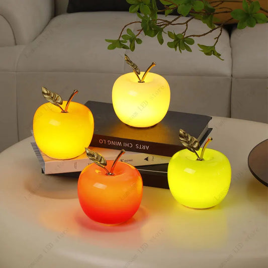 Four apple-shaped lamps in different colors on a table with books and a plant in the background.