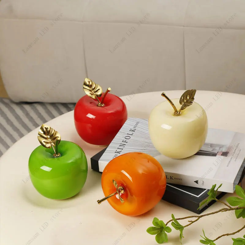 Four decorative apples in red, green, orange, and white on a surface with books underneath.