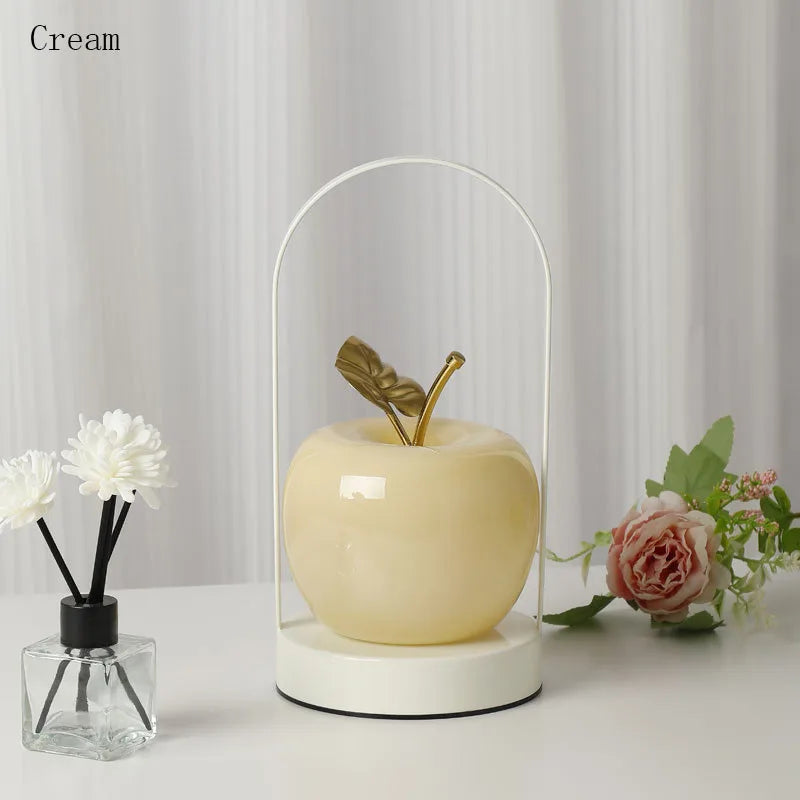 Decorative apple with a gold leaf inside a glass dome on a white surface with flowers and a diffuser.