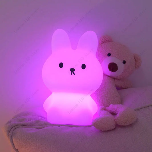 Cute rabbit-shaped night light emitting pink glow next to a teddy bear on a soft surface.