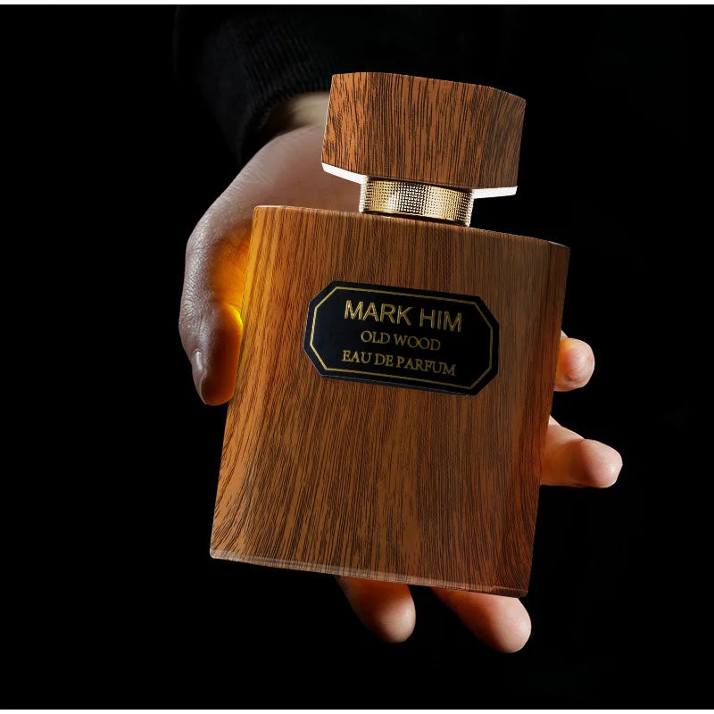 Wooden perfume bottle labeled 'Mark Him Old Wood Eau de Parfum' held by a hand against a black background