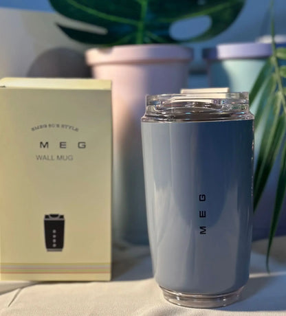 SMEG Silver tumbler with 'MEG' branding next to its packaging on a surface with plants in the background.