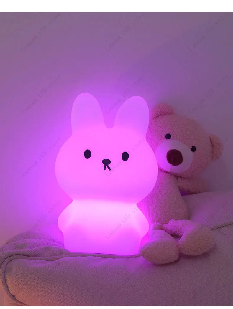 Cute rabbit-shaped night light emitting a pink glow, sitting next to a teddy bear on a soft surface.