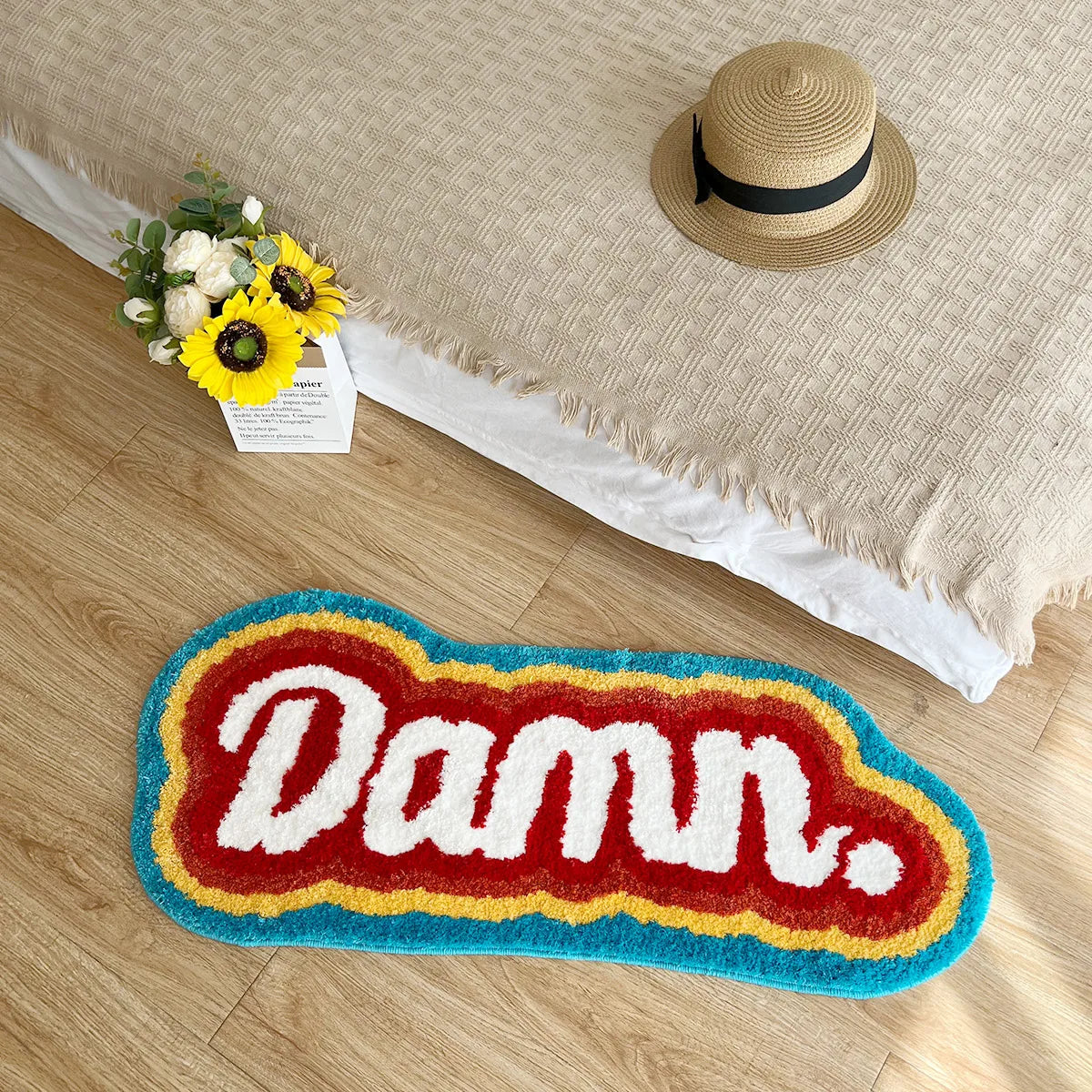 Decorative doormat with 'Damn.' text on a wooden floor, next to a hat and flowers.